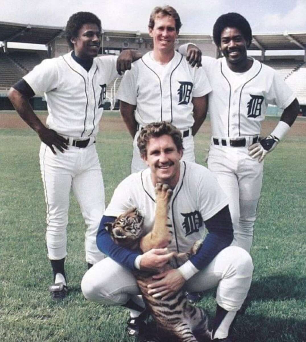 Four Detroit Tigers baseball players pose on a field; three stand in the background while one kneels in front, holding a playful tiger cub. The stadium seats are visible in the background.