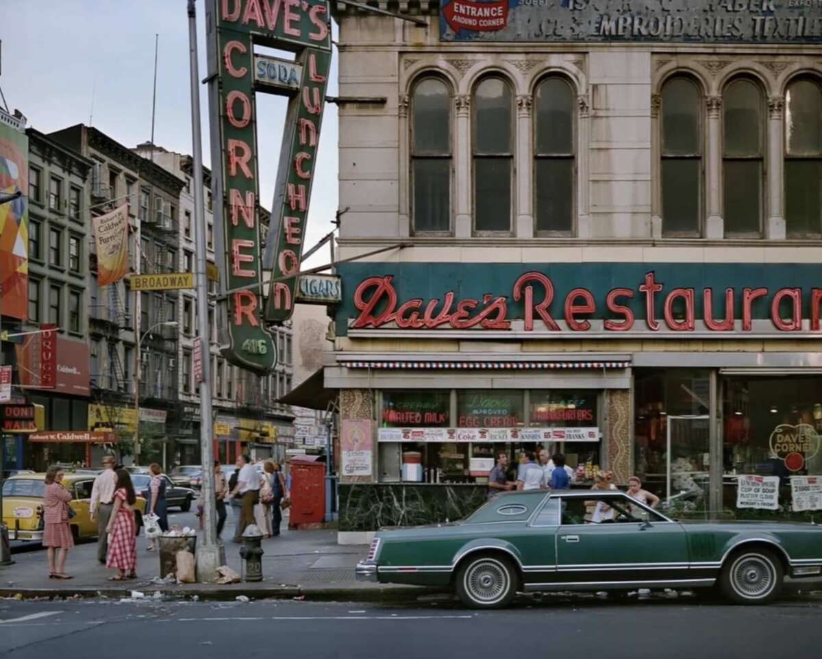 A street scene shows people walking past Dave’s Restaurant, a vintage diner with neon signs, large windows, and an old green car parked in front. The surrounding buildings have retro storefronts and colorful signs.
