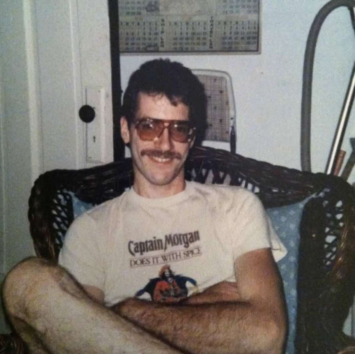 A man with curly hair, a mustache, and sunglasses sits cross-legged in a wicker chair. He wears a "Captain Morgan" t-shirt and smiles at the camera. A calendar hangs on the wall behind him.