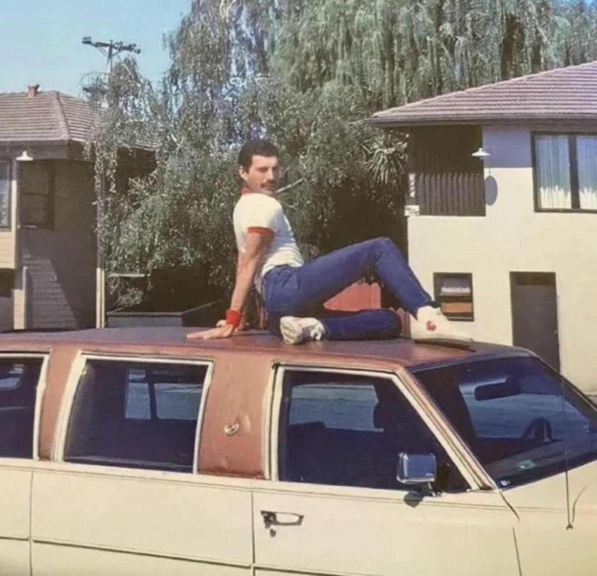 A man in a white shirt and blue jeans poses sitting on the roof of a brown car, with houses and trees in the background on a sunny day.