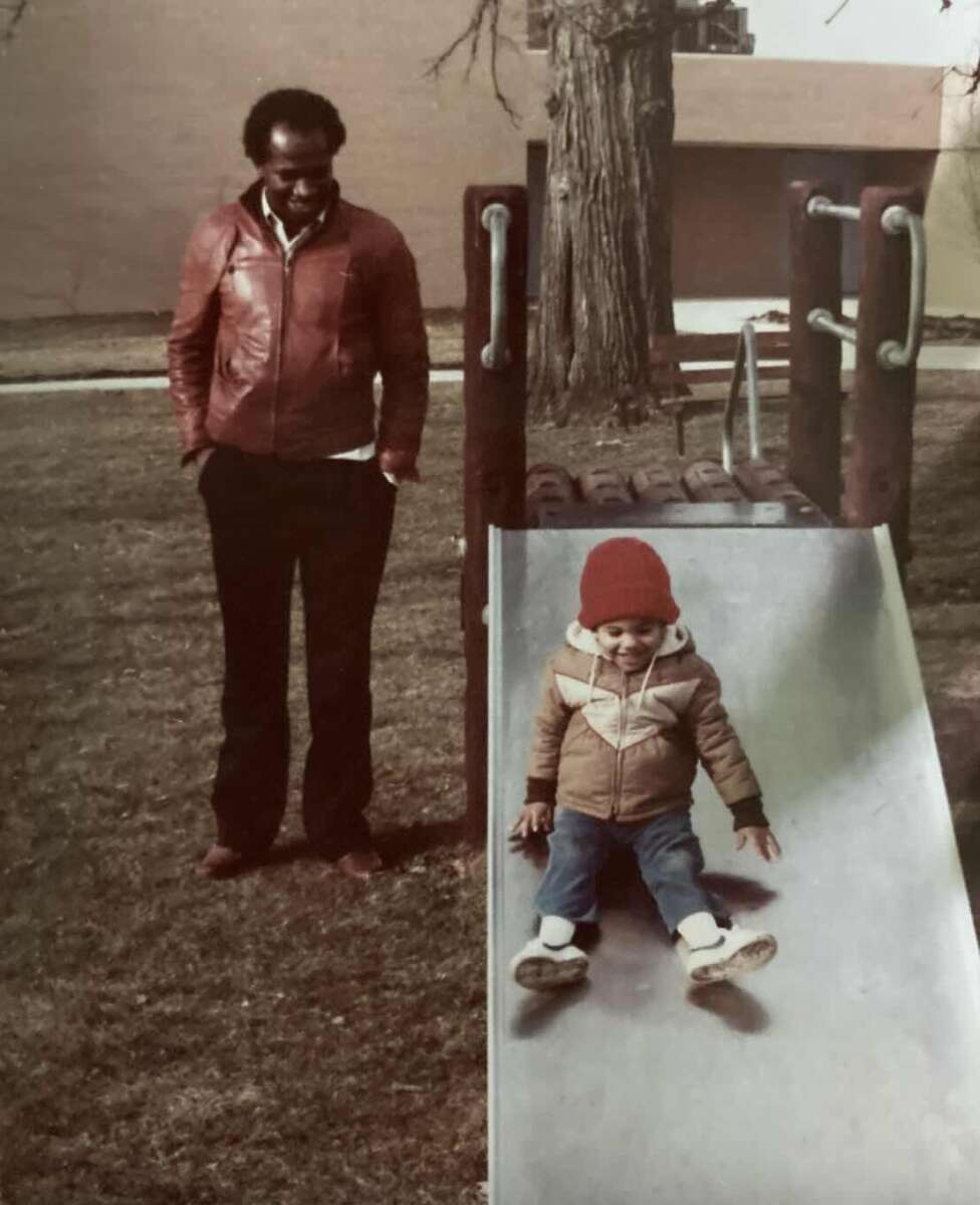A man in a red jacket stands beside a playground slide, watching a child in a red hat and winter coat slide down. The scene takes place in a grassy park with a large tree in the background.