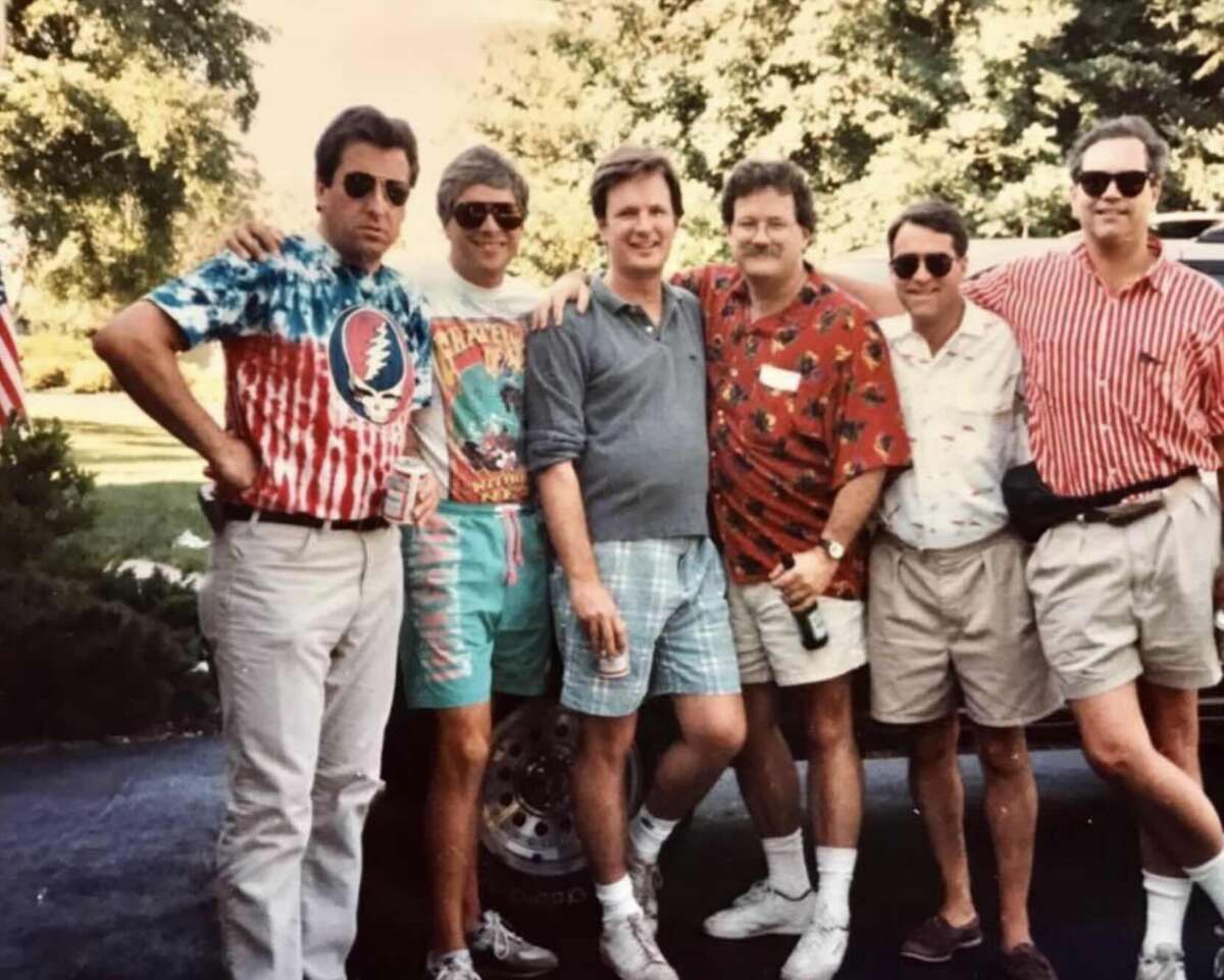 Six men stand outdoors in casual summer clothing, smiling for the camera. They are wearing colorful shirts and shorts, with trees and a car in the background. The mood is relaxed and friendly.