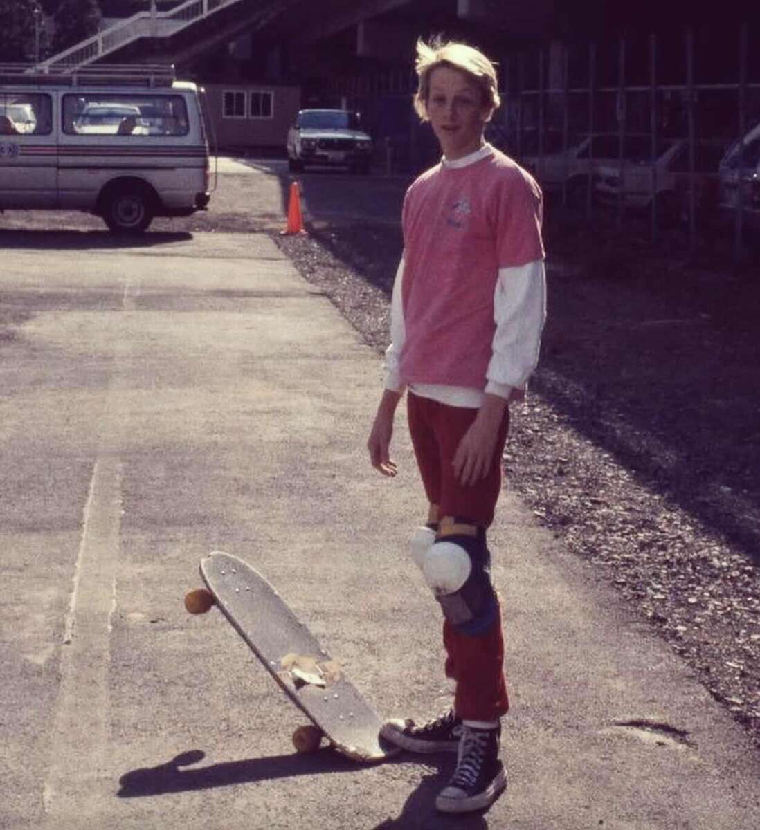 A young person stands outdoors on a pavement with a skateboard tilted beside their feet, wearing a pink shirt over a white long-sleeve, red pants, knee pads, and sneakers. A van and building are in the background.