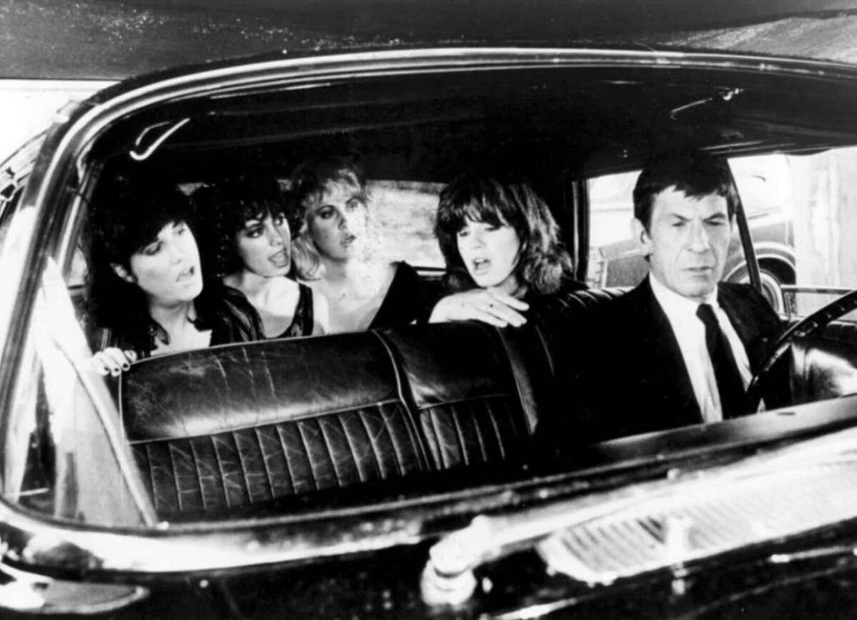 Four women sit in the backseat of a car, while a man in a suit drives. The women appear to be talking animatedly, and the driver looks serious and focused on the road. The photo is in black and white.
