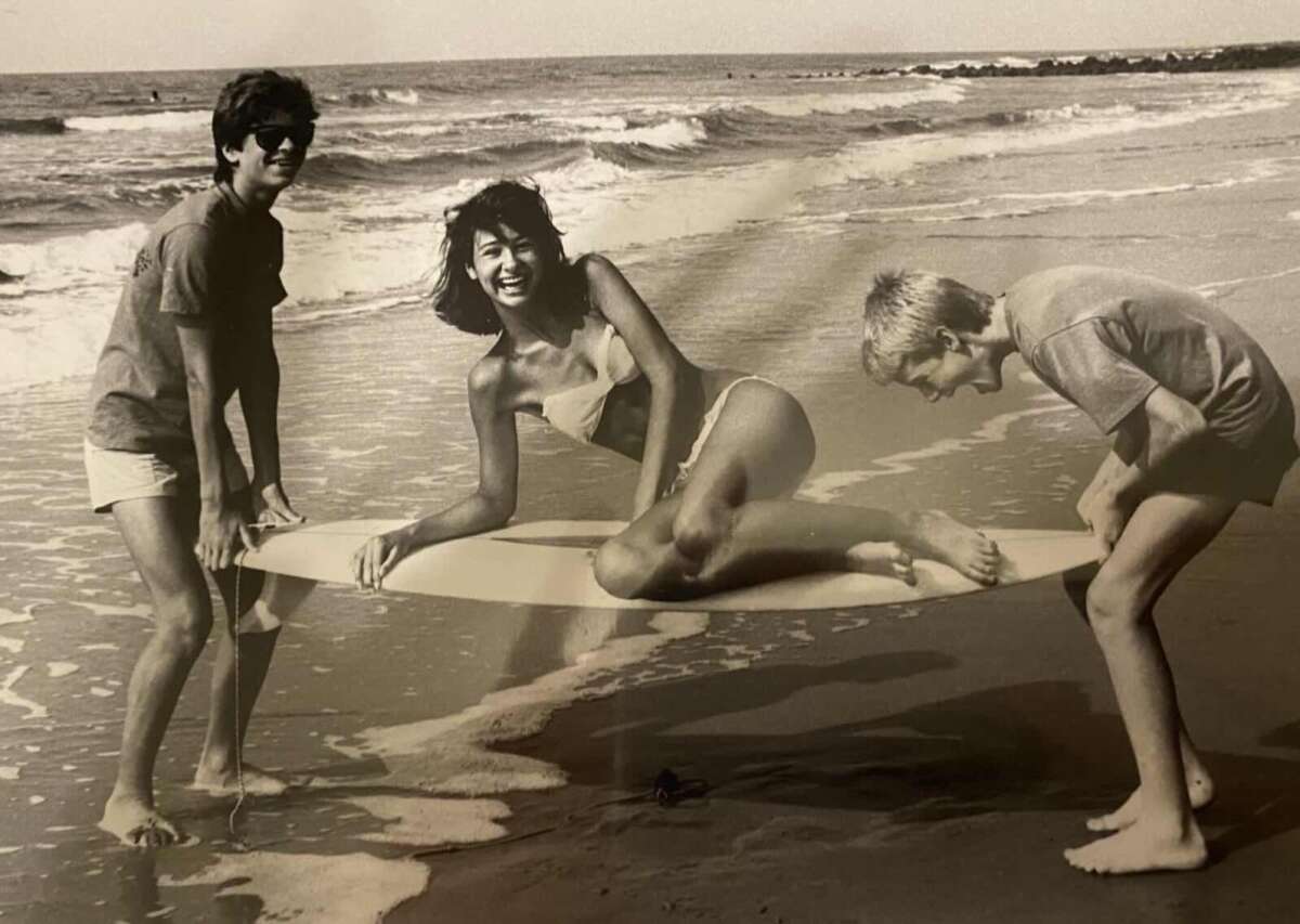 Three people on a beach hold a surfboard above the wet sand and water; a woman in a bikini sits smiling on the board, while two boys in shorts hold either end, all appear happy. The photo is in black and white.