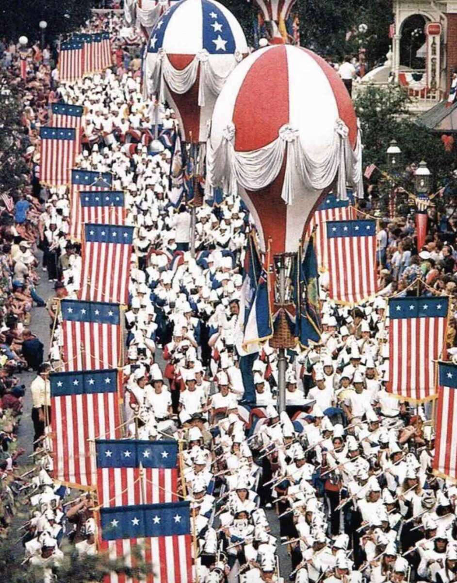 A parade with people dressed in white uniforms marching between large American flags and two tall red, white, and blue balloon floats, surrounded by a crowd of spectators.