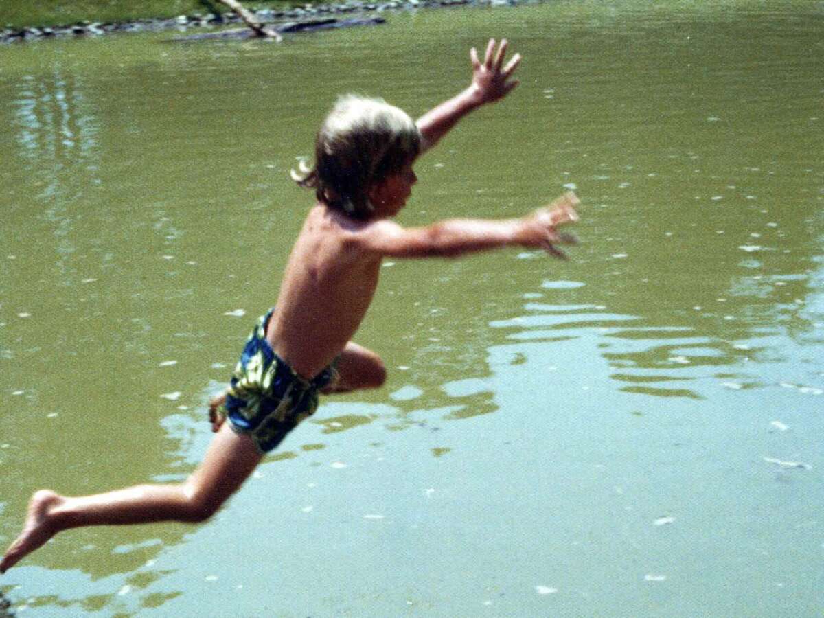 A young child in swim trunks leaps with arms outstretched toward a pond, mid-air above the water on a sunny day.