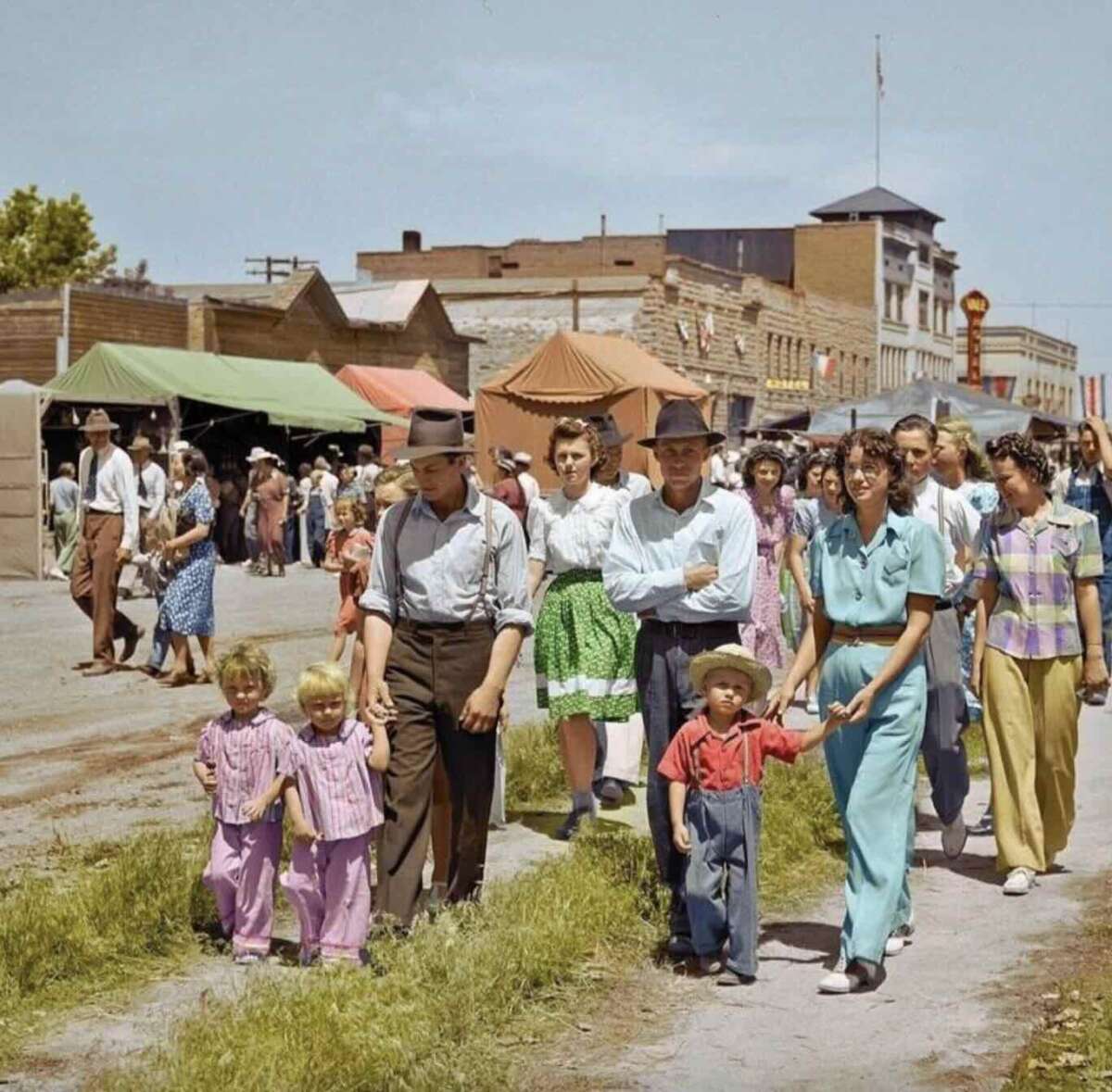 A group of people, including adults and children in colorful 1940s-style clothing, walk together at an outdoor fair or market on a sunny day with tents, shops, and crowds in the background.