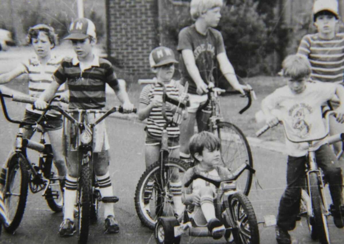 A black and white photo of seven children outdoors, most on bicycles or tricycles. They wear striped shirts, shorts, and caps, appearing to be ready for a bike ride. The background shows trees and a building.