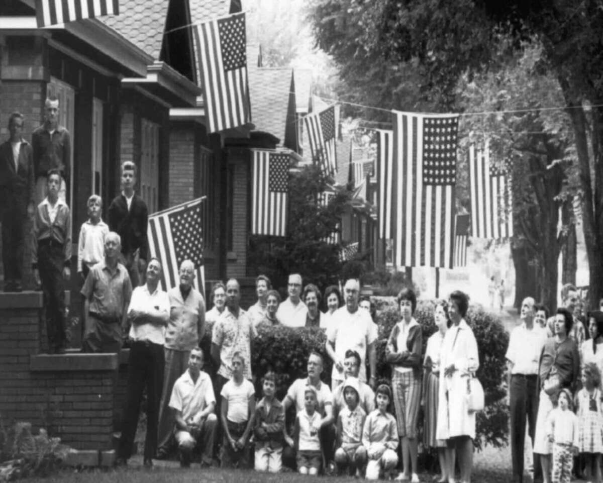 A black-and-white photo shows a large group of people, including children and adults, gathered outside suburban homes decorated with numerous American flags hanging along the street.