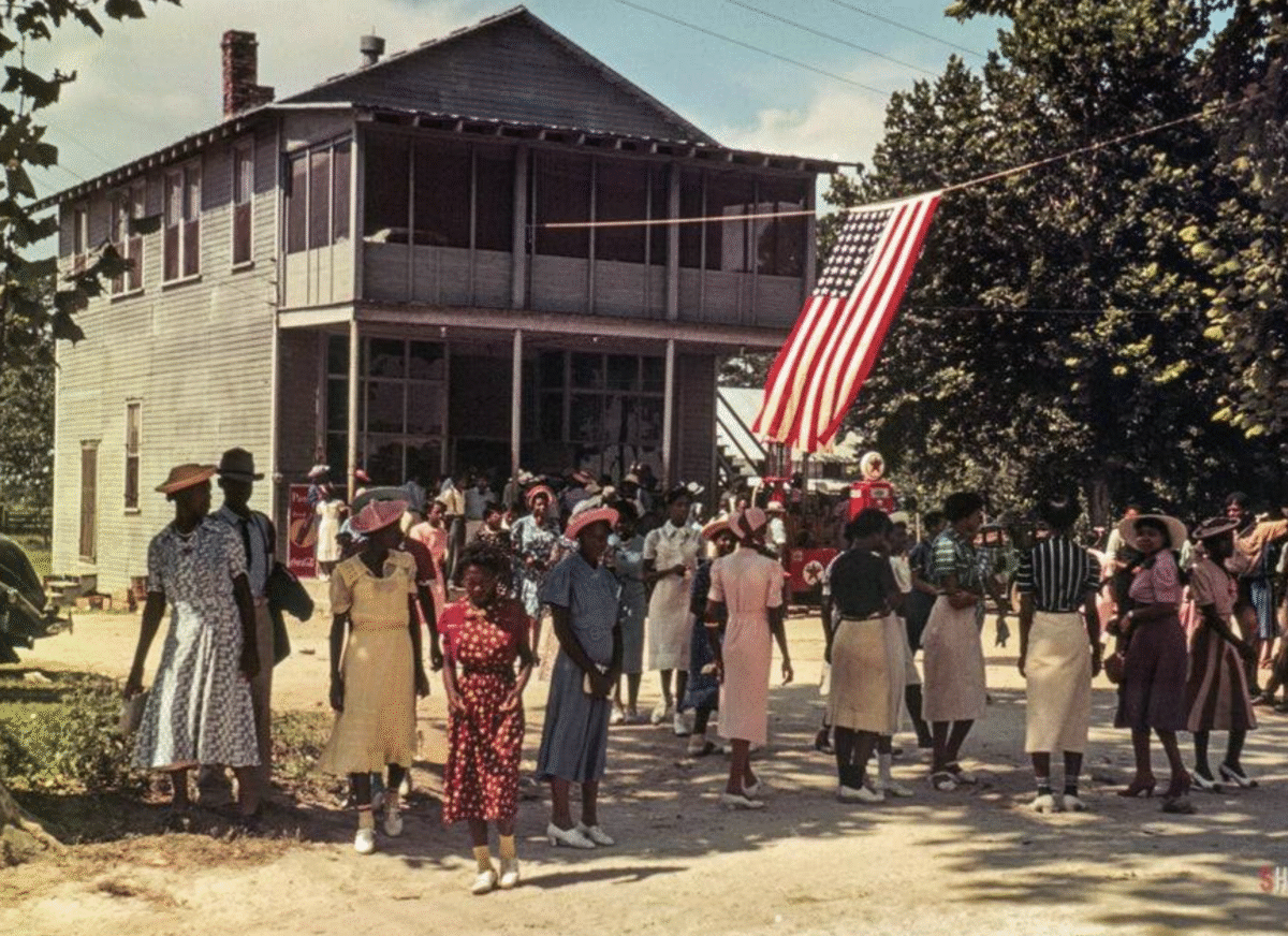 A group of people, mostly women and girls in colorful dresses and hats, gather outside a large wooden house with an American flag hanging overhead on a sunny day. Trees frame the scene.