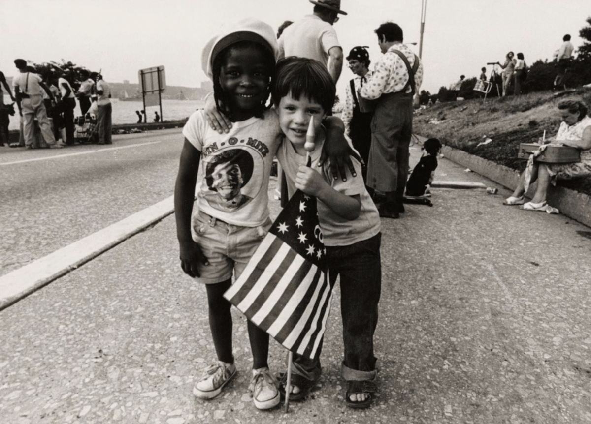 Two children, one Black and one white, smile and hug each other while holding an American flag on a street. People and photographers stand in the background, and a woman sits on a curb.