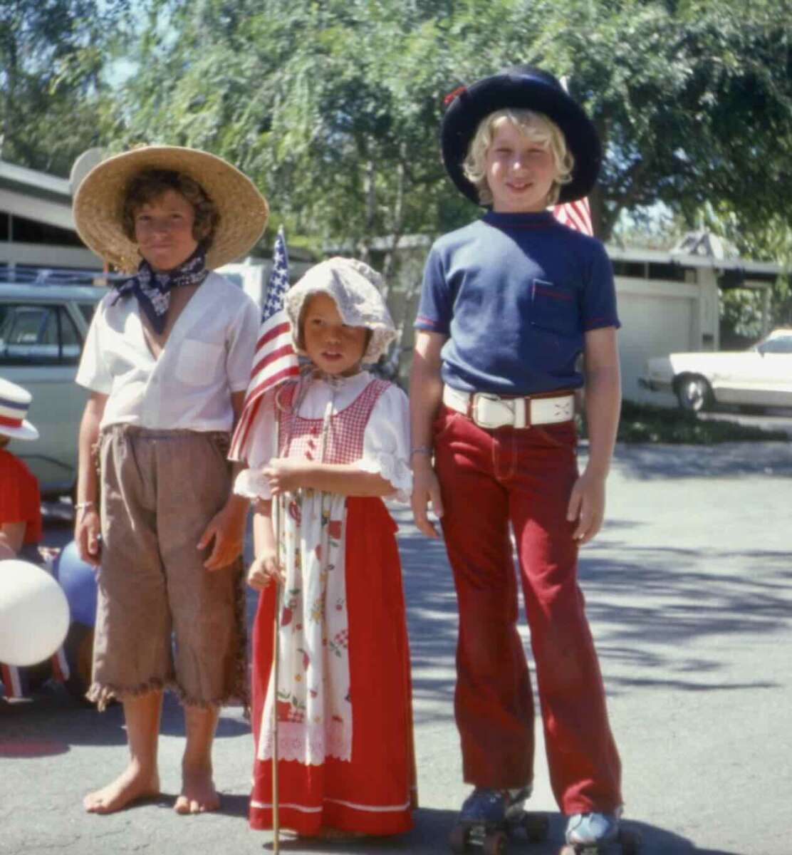 Three children in vintage-style clothing stand outdoors. One wears tattered pants and a straw hat, another is in a red dress and bonnet holding an American flag, and the third wears red pants and a blue hat. Cars and trees are in the background.