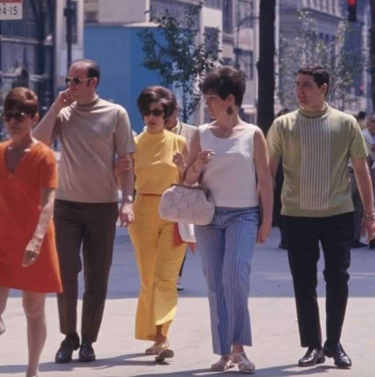 Five people in colorful 1960s-style clothing walk down a city street. Three women wear bright dresses or tops and two men wear short-sleeved shirts and trousers. Buildings and trees line the sidewalk in the background.