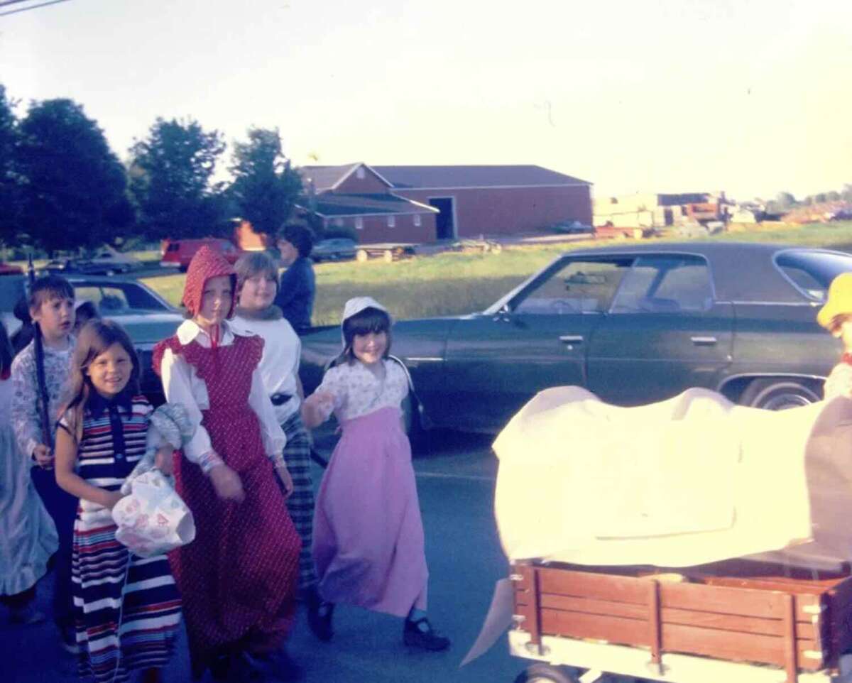 A group of children in vintage-style clothing walk outside on a sunny day, smiling. Behind them are old cars, a grassy area, and barns or large buildings in the distance. A red wagon is partially visible in the foreground.