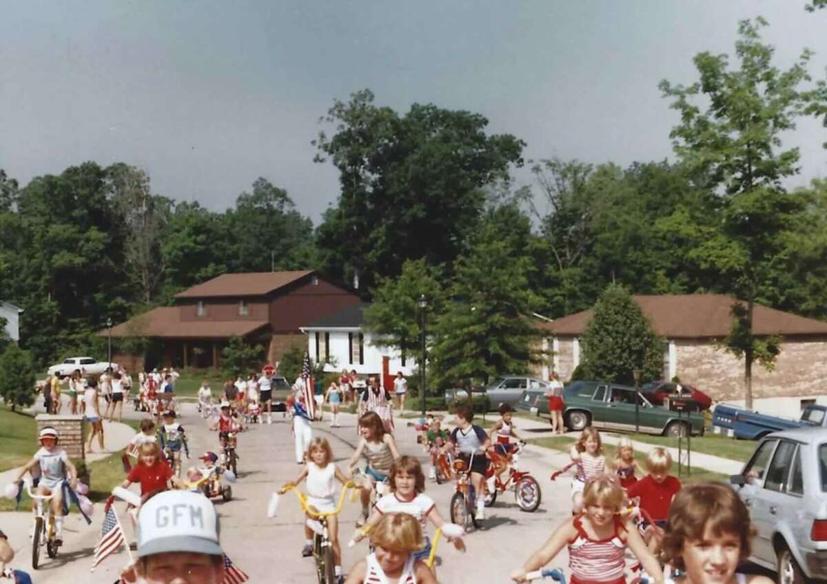 Children ride decorated bicycles and tricycles down a suburban street during a parade, with adults watching and houses and trees in the background. Many wear red, white, and blue outfits, suggesting a patriotic celebration.