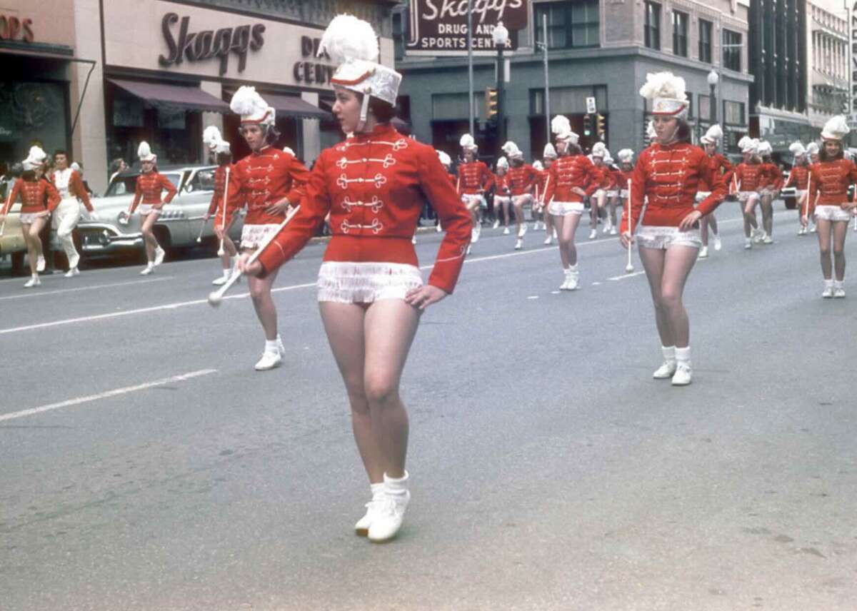A group of female majorettes in red and white uniforms and white hats march down a city street during a parade, with storefronts and parked cars visible in the background.