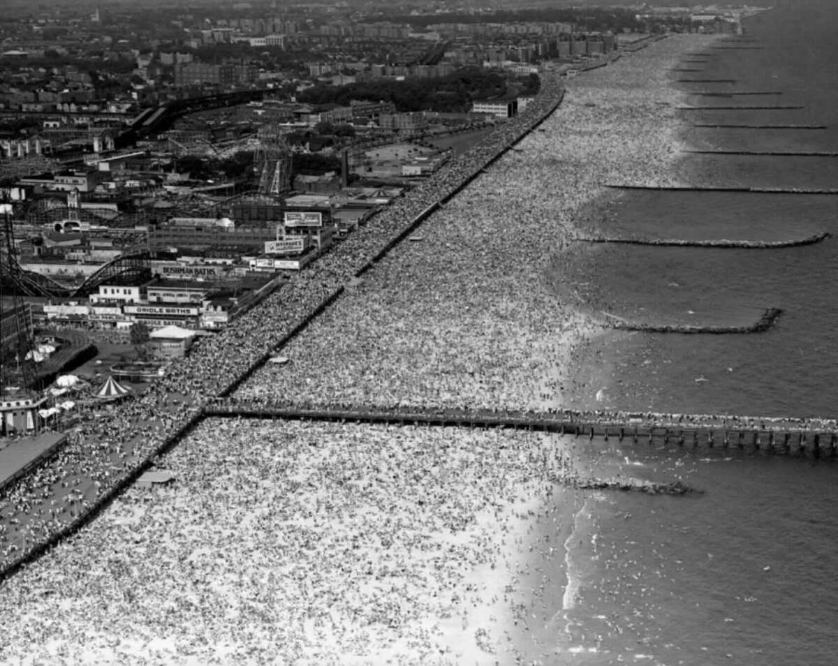 Aerial black-and-white photo of a crowded beach next to a city, with thousands of people gathered along the shoreline and boardwalk, and piers extending into the ocean.