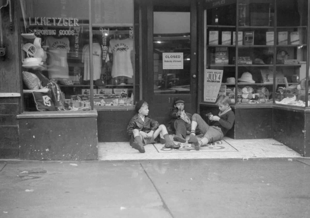 Three boys sit on a sidewalk in front of a closed shop, talking and relaxing. The storefront displays sporting goods and hats, and a "Closed" sign hangs on the door. The street looks wet from recent rain.