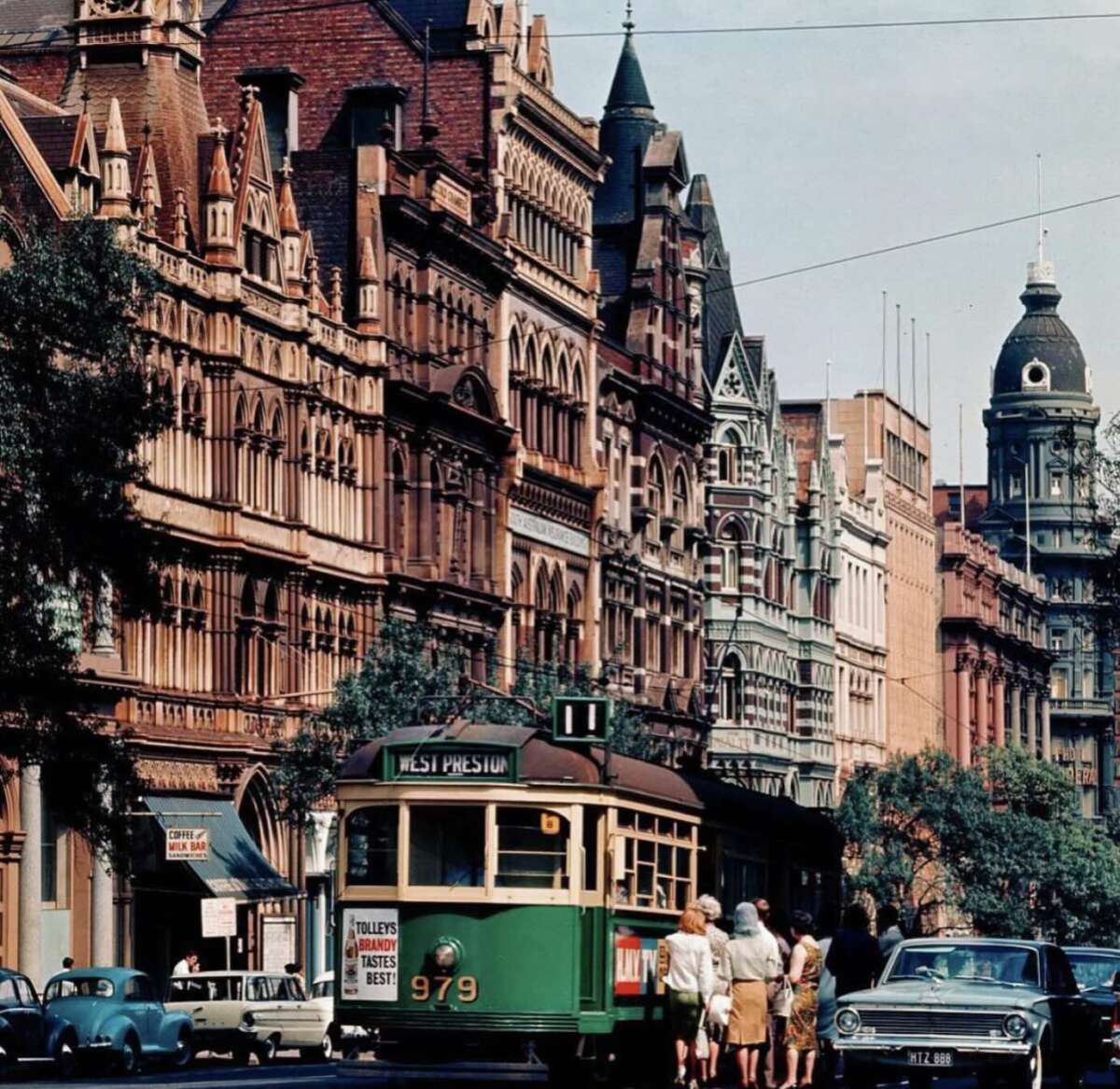 A vintage green tram with passengers boarding in front of ornate, historic buildings on a lively city street lined with parked cars and trees.