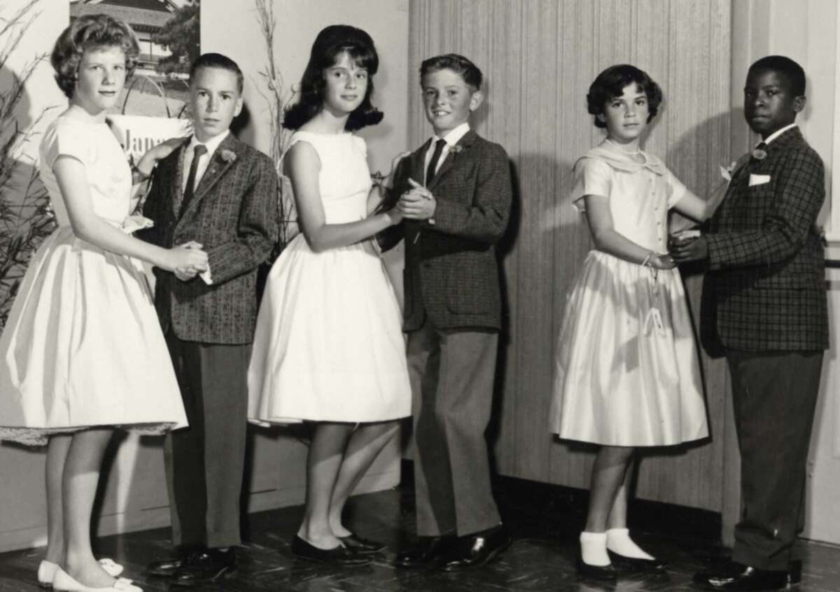 Six children dressed formally, standing in pairs and dancing. The boys wear suits and the girls wear dresses. They are inside, posing in a line, with a wooden wall behind them. The image is black and white.