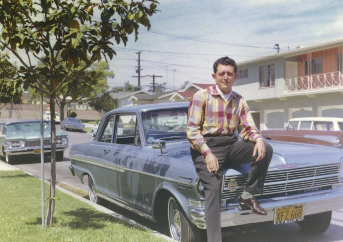 A man in a plaid shirt and dark pants sits on the hood of a vintage car parked on a suburban street, with houses and another classic car visible in the background.