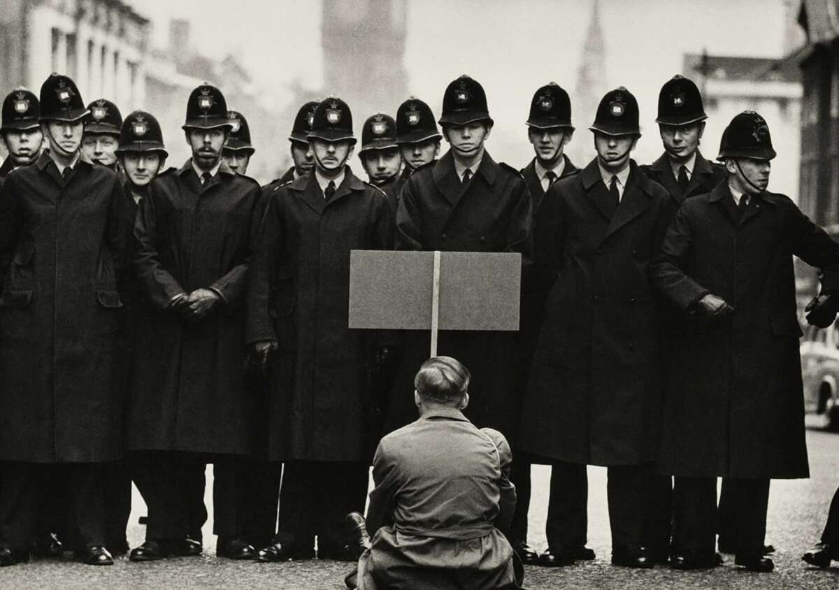 A lone protester sits on the street facing a group of British police officers standing in a row, blocking the road. The protester holds a sign, and the officers wear long coats and helmets. The scene appears tense and serious.