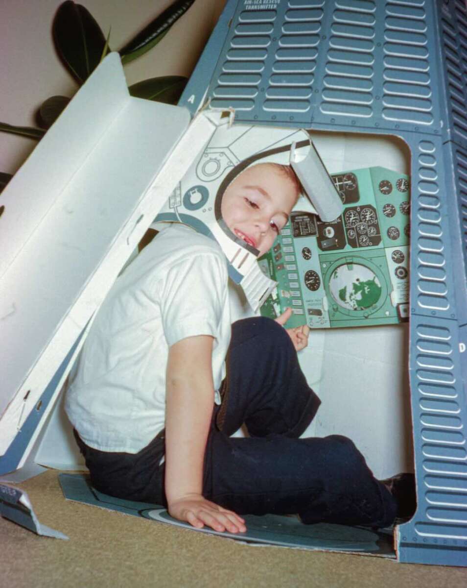 Young child sits inside a cardboard play rocket with illustrated control panels, wearing a paper astronaut helmet and smiling. The scene evokes imaginative play and space exploration.