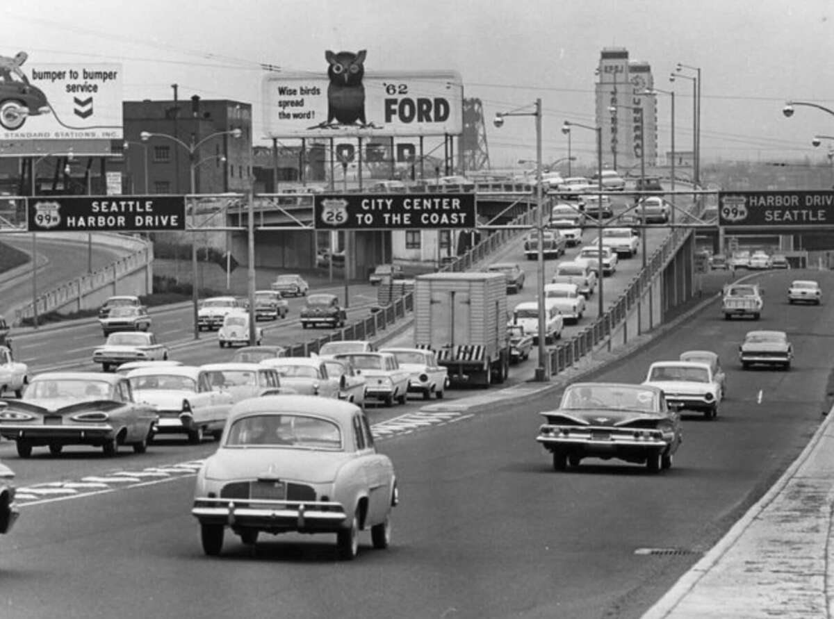 Black and white photo of heavy traffic on a multi-lane highway in Seattle, with 1960s-era cars, large road signs, and city buildings in the background. A billboard advertises Ford and a bird food brand.