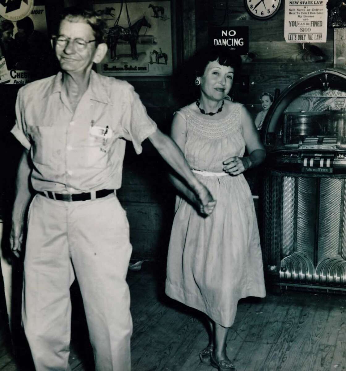 An older man and woman hold hands while dancing in a vintage bar, with a jukebox, a “No Dancing” sign, and retro posters visible in the background.