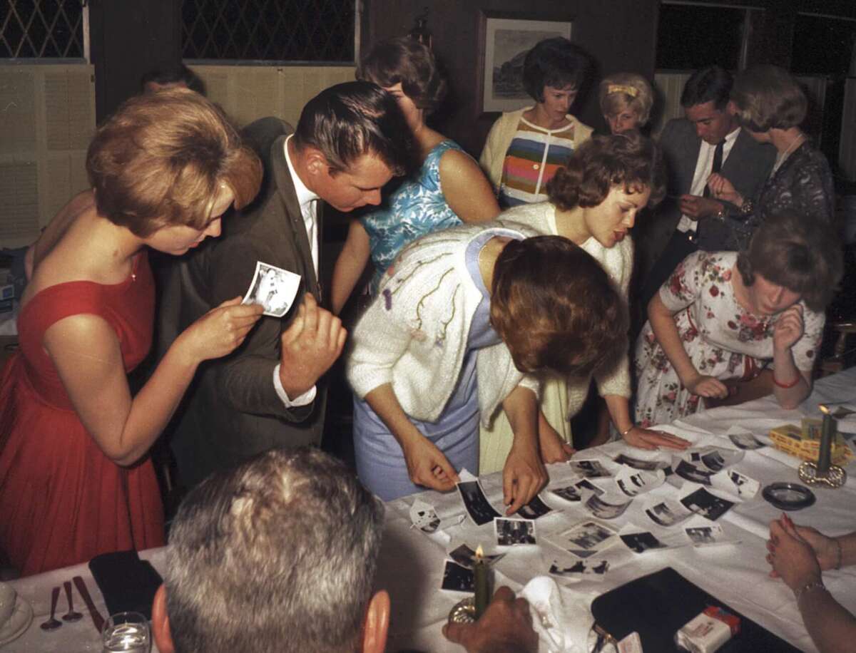 A group of people in formal attire gather around a table, closely examining black-and-white photographs spread out on a white tablecloth during an indoor social event.
