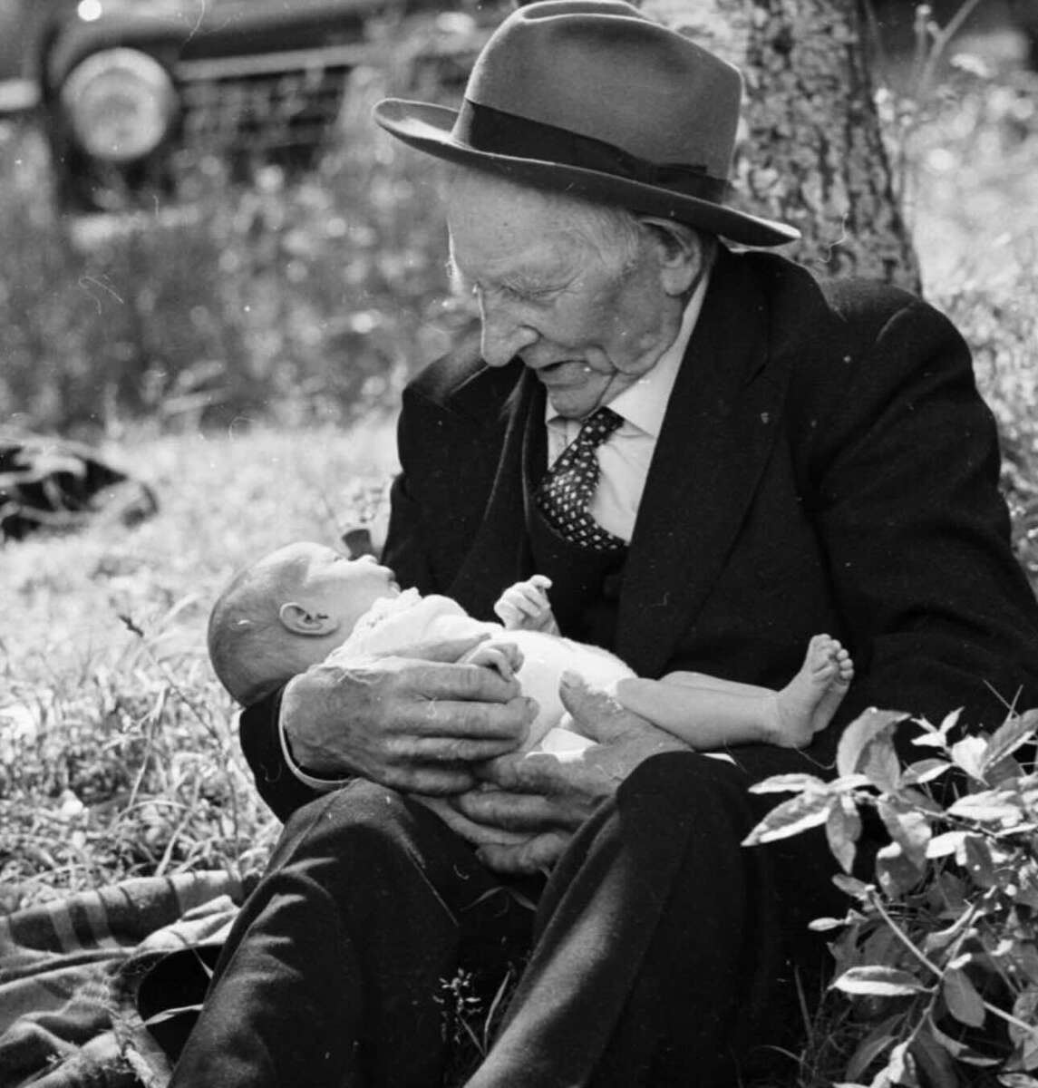 An elderly man in a suit and hat sits on the grass, cradling a baby in his arms and smiling warmly at the child on a sunny day. A vintage car and trees are visible in the blurred background.