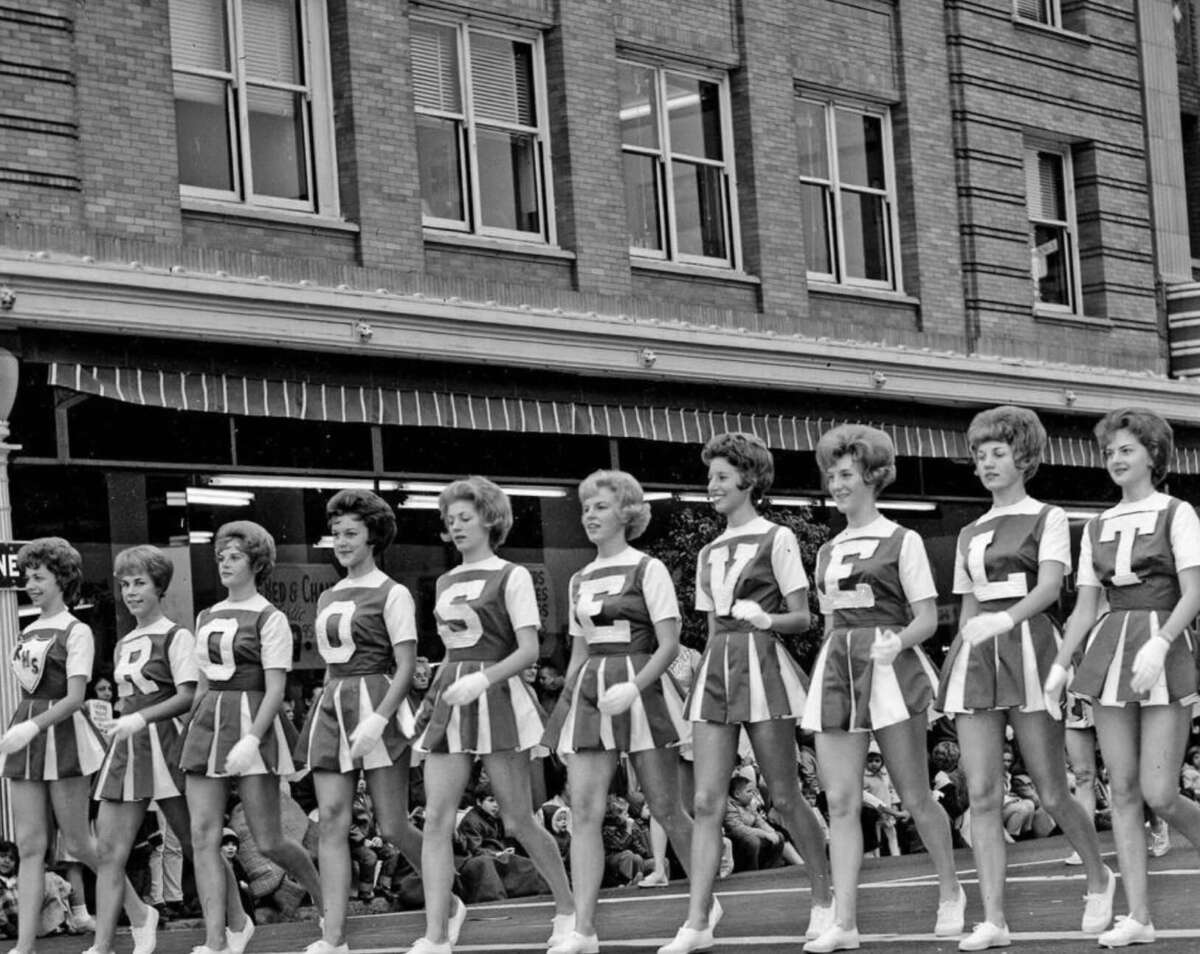 A group of cheerleaders in matching uniforms stand in a row on a parade float, each with a letter on their top spelling "ROOSEVELT," with a brick building and crowd in the background.