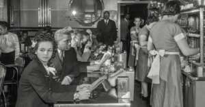 A crowded diner in the 1950s with Black and white patrons sitting at the counter, reading menus, while waitresses in uniforms serve food and drinks. The atmosphere is busy, with people engaged in conversation.