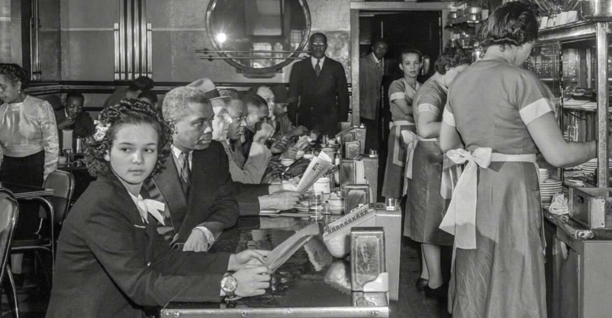 A crowded diner in the 1950s with Black and white patrons sitting at the counter, reading menus, while waitresses in uniforms serve food and drinks. The atmosphere is busy, with people engaged in conversation.
