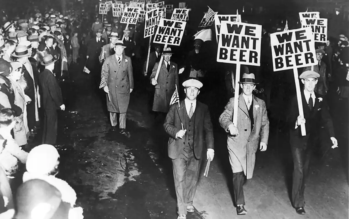 A group of men in suits and hats march in a nighttime protest, holding signs that read "WE WANT BEER." The crowd appears determined, and some people in the background are watching the demonstration.