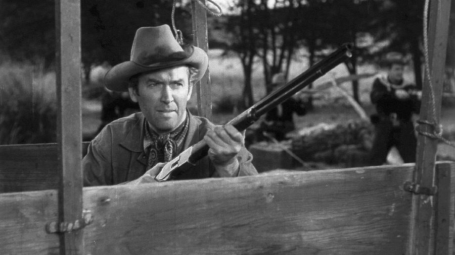 A man in a cowboy hat aims a rifle while hiding behind a wooden barrier outdoors; blurred figures in the background also appear armed. The scene is in black and white.