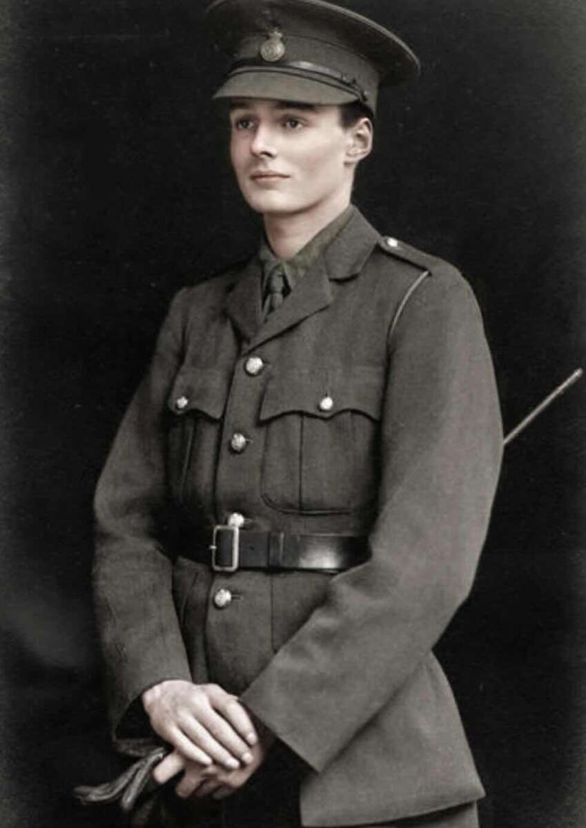 A young person in a vintage military uniform and cap stands against a dark background, hands clasped in front, looking slightly to the side with a calm expression.