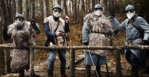 Four soldiers in vintage uniforms, fur vests, and helmets stand alert on a wooden bridge in a forest, wearing protective masks over their faces. The atmosphere appears tense and wintry.