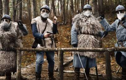 Four soldiers in vintage uniforms, fur vests, and helmets stand alert on a wooden bridge in a forest, wearing protective masks over their faces. The atmosphere appears tense and wintry.