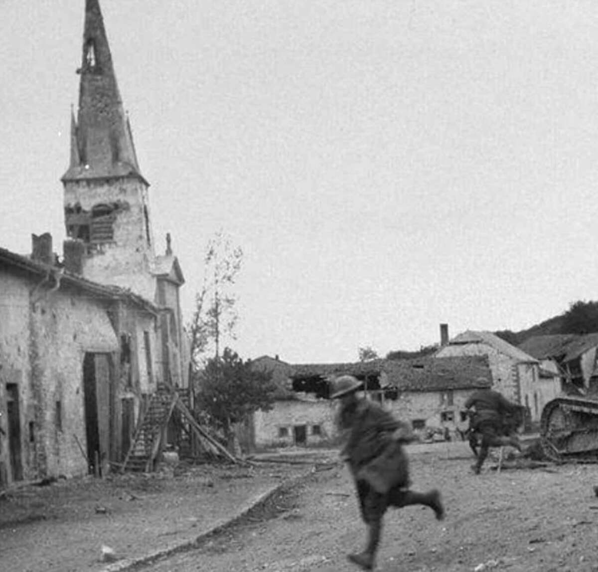 A black-and-white photo shows two soldiers running through a war-torn village street with damaged buildings and a church with a broken steeple in the background. Debris and an armored vehicle are visible.