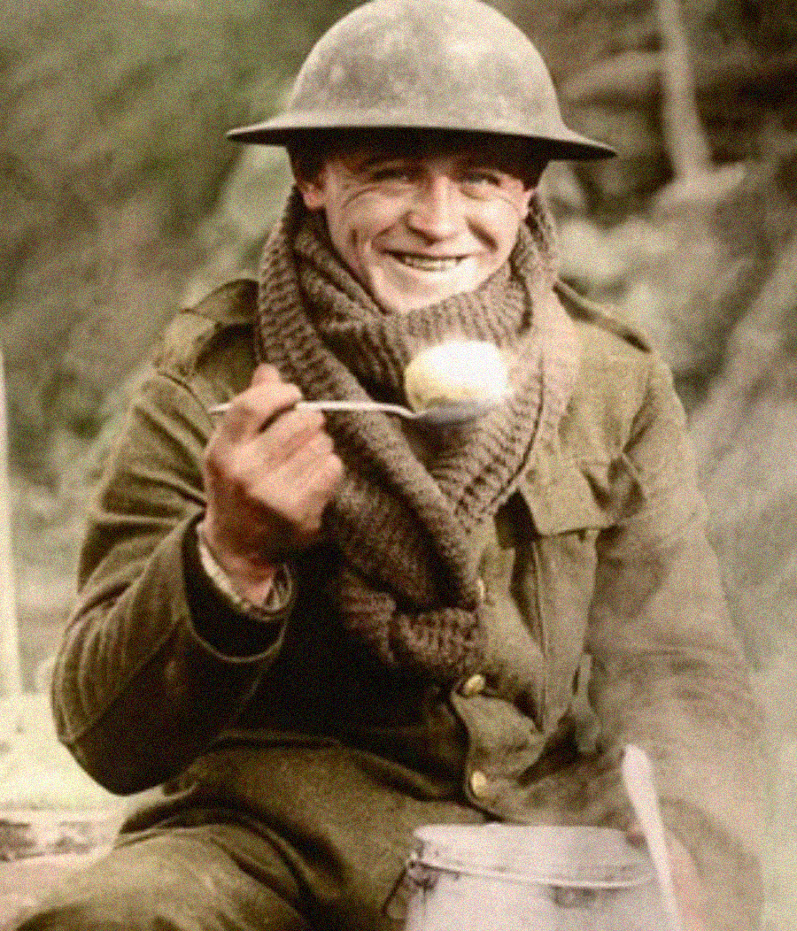 A smiling soldier in a helmet and scarf holds up a spoon with food, sitting outdoors next to a metal pot. The image has a vintage, historical appearance.