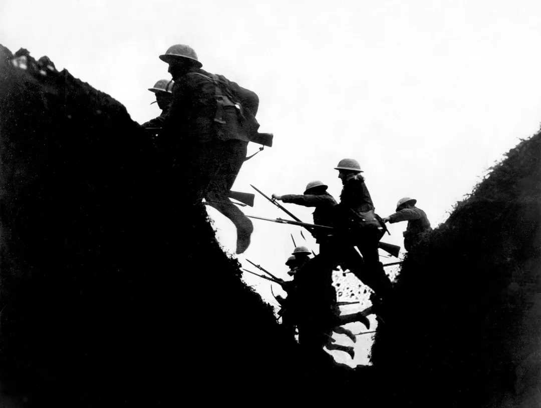 Silhouetted soldiers in helmets and uniforms, armed with rifles and bayonets, climb out of a trench during wartime, moving upward against a bright, overexposed sky.
