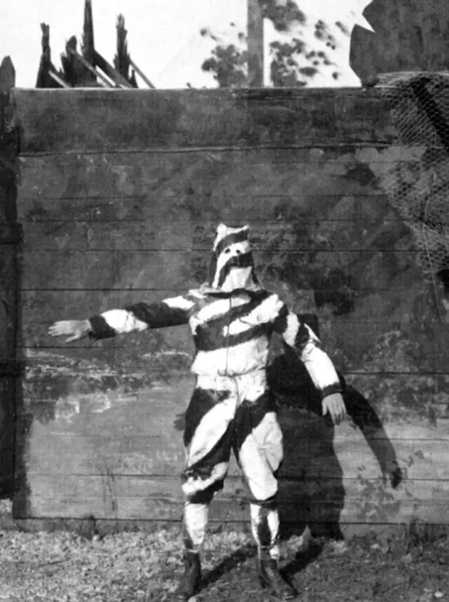 A person stands with arms outstretched, wearing a full-body suit with bold, irregular black and white camouflage patterns, in front of a wooden wall outdoors.