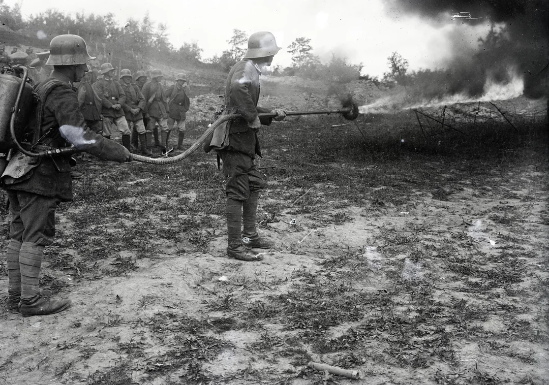 Several soldiers in World War I-era uniforms observe as one soldier uses a flamethrower, producing a spray of fire and smoke outdoors on a patch of bare earth with trees in the background.