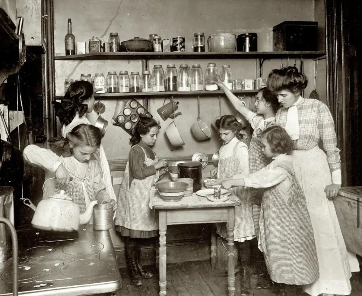 A group of six girls in old-fashioned dresses and aprons work together in a vintage kitchen, pouring, mixing, and preparing food around a wooden table with shelves of jars and dishes behind them.