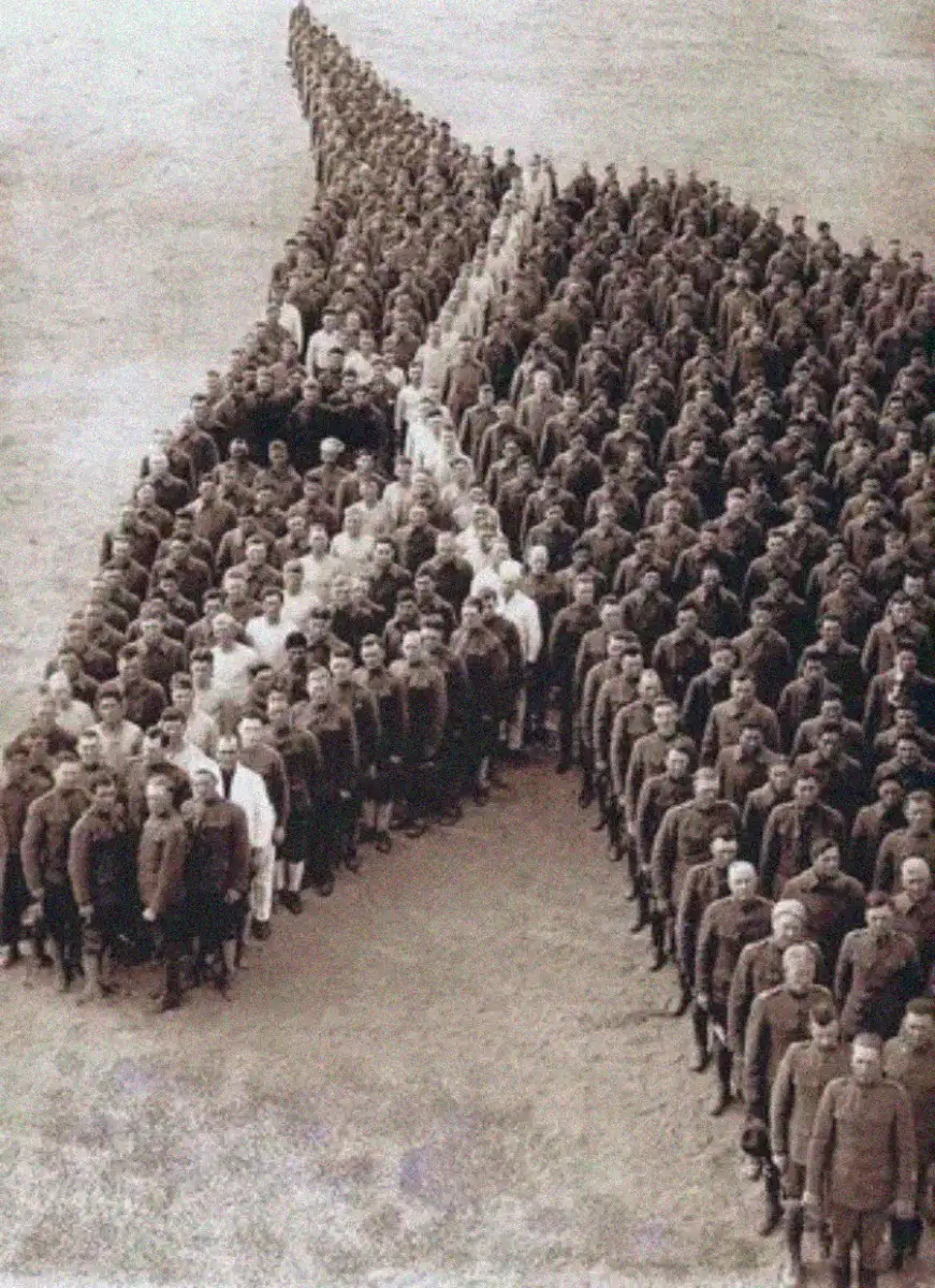 A large group of people standing together in formation, creating the shape of an arrow with a hollow center, in a sepia-toned photograph taken outdoors.