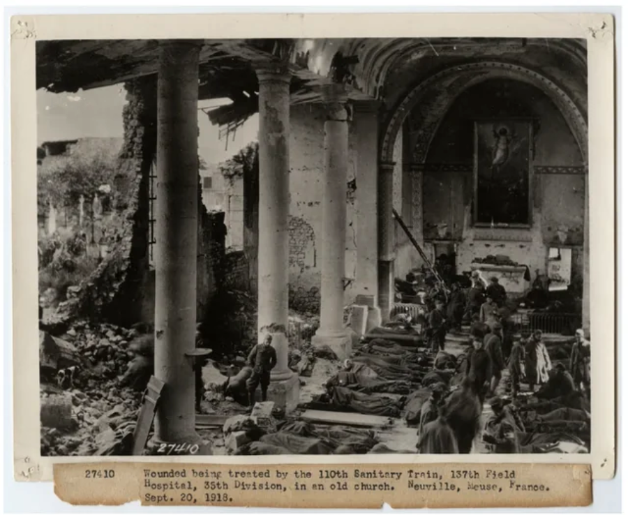 Black and white photo of wounded soldiers lying on cots inside a damaged church, with medics attending to them. Debris and broken columns are visible. Caption notes it's a field hospital in Neuville, France, 1918.