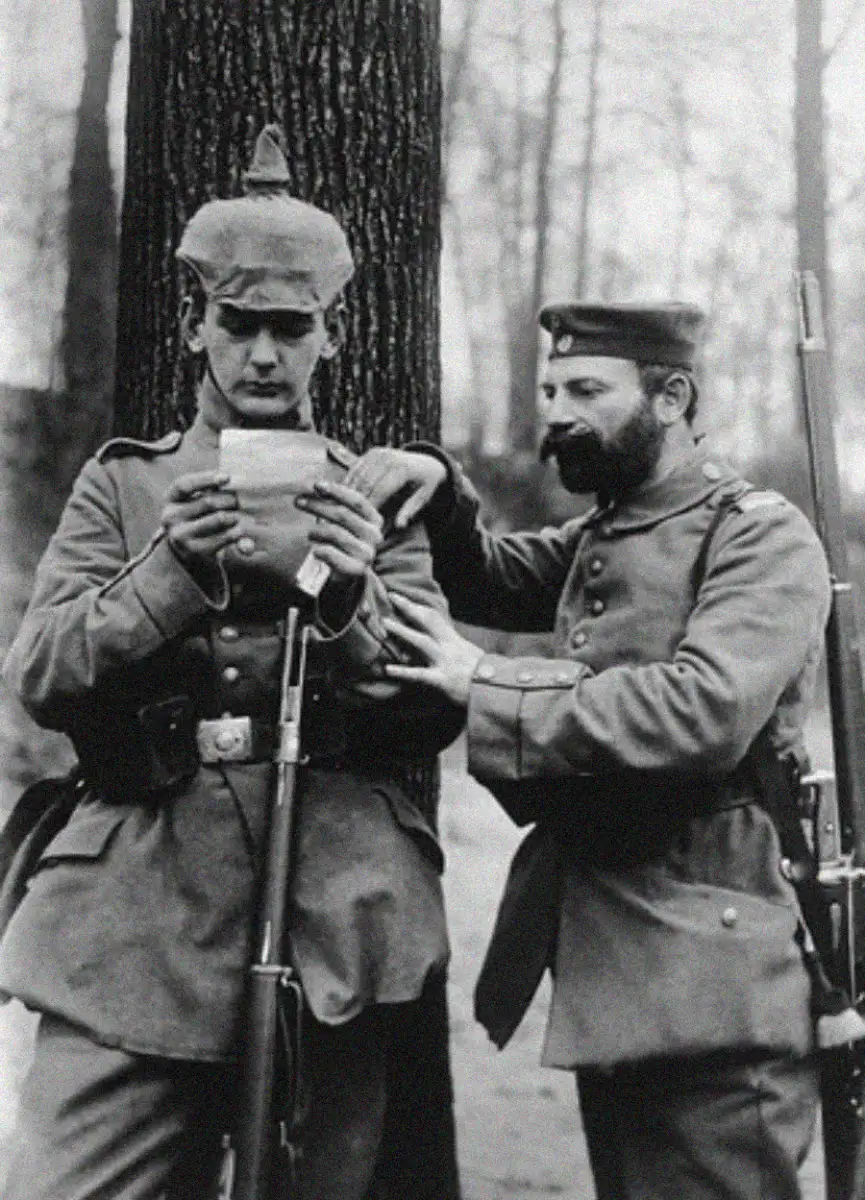 Two soldiers in early 20th-century military uniforms stand by a tree. One soldier reads a letter while the other looks on, resting his arm gently on his comrade's shoulder. Both have rifles and serious expressions.