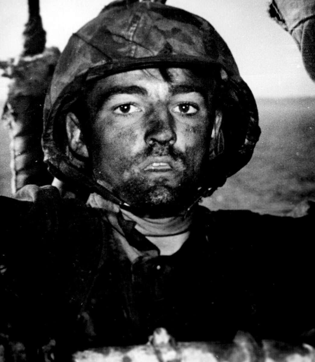 A close-up black and white photo of a soldier in a helmet, with a dirty face, gazing directly at the camera. The background is blurred, suggesting an outdoor setting.